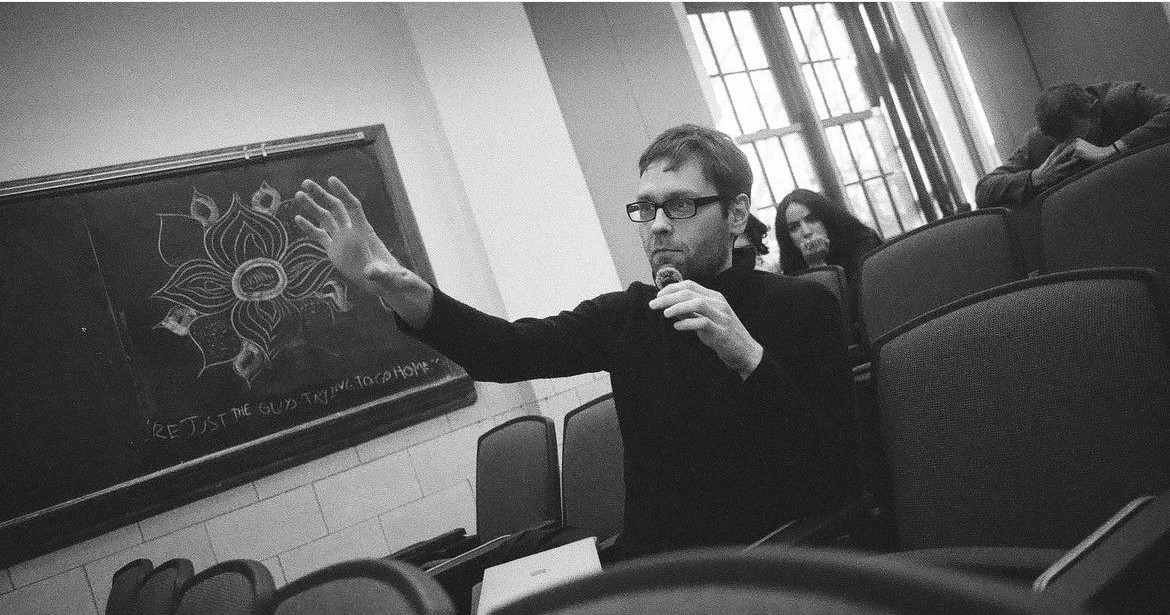 Robert Reimer raising his hand to ask a question during a philosophy talk, with a chalk drawing on the blackboard behind him.